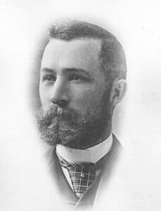 A black and white portrait of a late 19th-century man with a beard and formal attire.