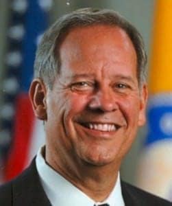 A smiling middle-aged man in professional attire at The History Museum, with colorful flags in the background.