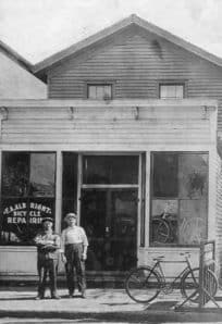 Faded black and white photo of a historic building with bicycles and two boys standing outside, early 20th century scene, The History Museum.