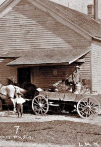 Horse-drawn milk delivery cart outside a wooden building in 19th-century rural America.