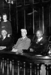 Historic photograph of distinguished guests and officials seated in a courtroom or council chamber, showcasing mid-20th century formal attire.