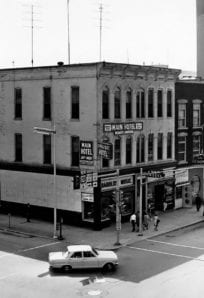 Old downtown building with Main Hotel, radio station and storefronts, vintage cars, and pedestrians.