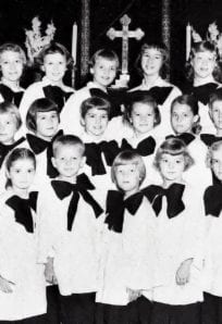 Children's choir dressed in white robes with black bows, standing in front of a church altar with a cross, from a historical church photo on the History Museum website.
