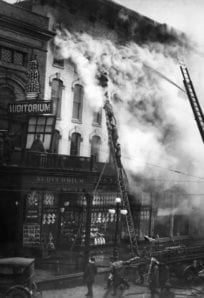 Fire damages the auditorium building, smoke billows from the top during an intense blaze in this historical black-and-white image.