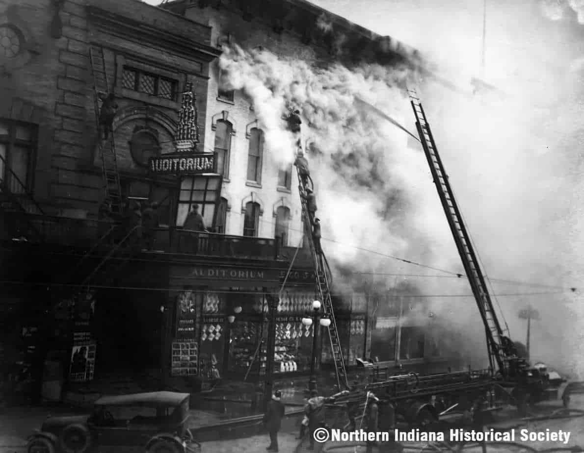 Fire damages the auditorium building, smoke billows from the top during an intense blaze in this historical black-and-white image.