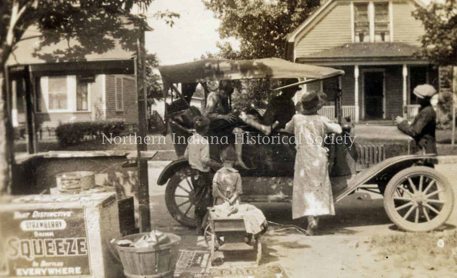 Vintage street scene with people around an early 20th-century car, historical houses, and ice cream vendor in Northern Indiana.