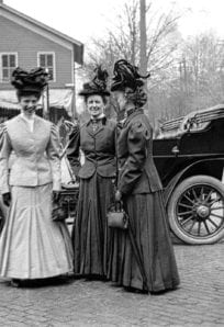 Women in early 20th century fashion standing in front of a vintage car outside the History Museum.