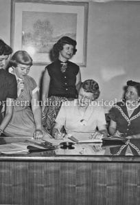 Women in 1950s office workspace discussing documents in a historic setting.