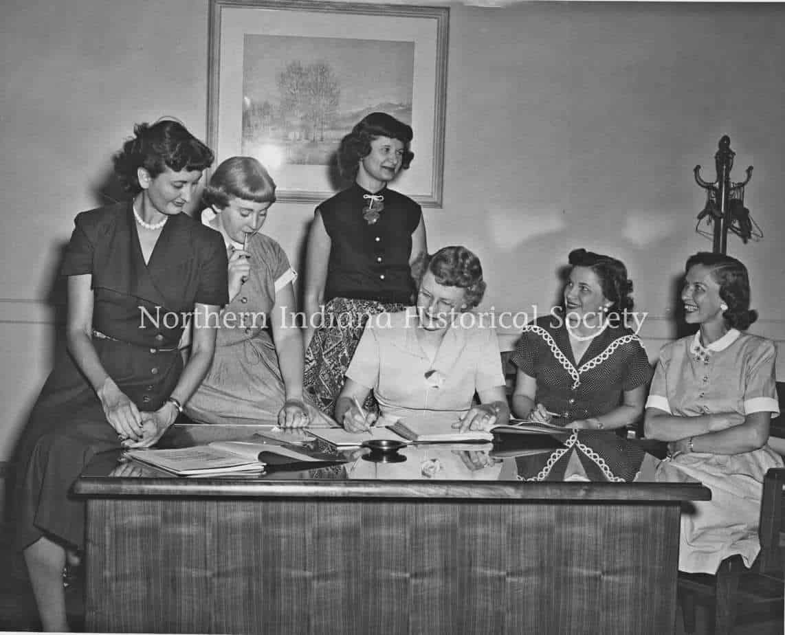 Women in 1950s office workspace discussing documents in a historic setting.