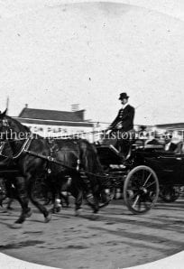 Horse-drawn carriage with people in vintage clothing, historic urban street scene, early 20th century.