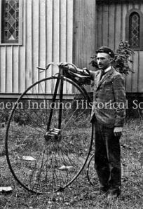 Winding wheel bicycle and young man outside wooden building, historical photo from Northern Indiana Historical Society, vintage outdoor scene.