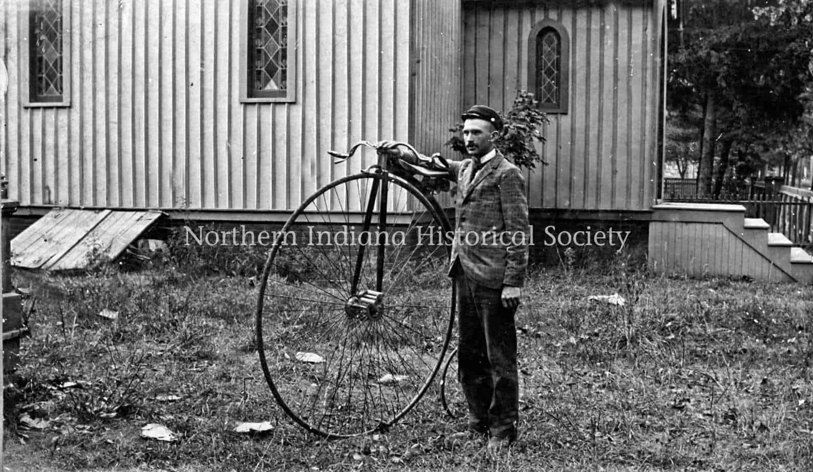 Winding wheel bicycle and young man outside wooden building, historical photo from Northern Indiana Historical Society, vintage outdoor scene.