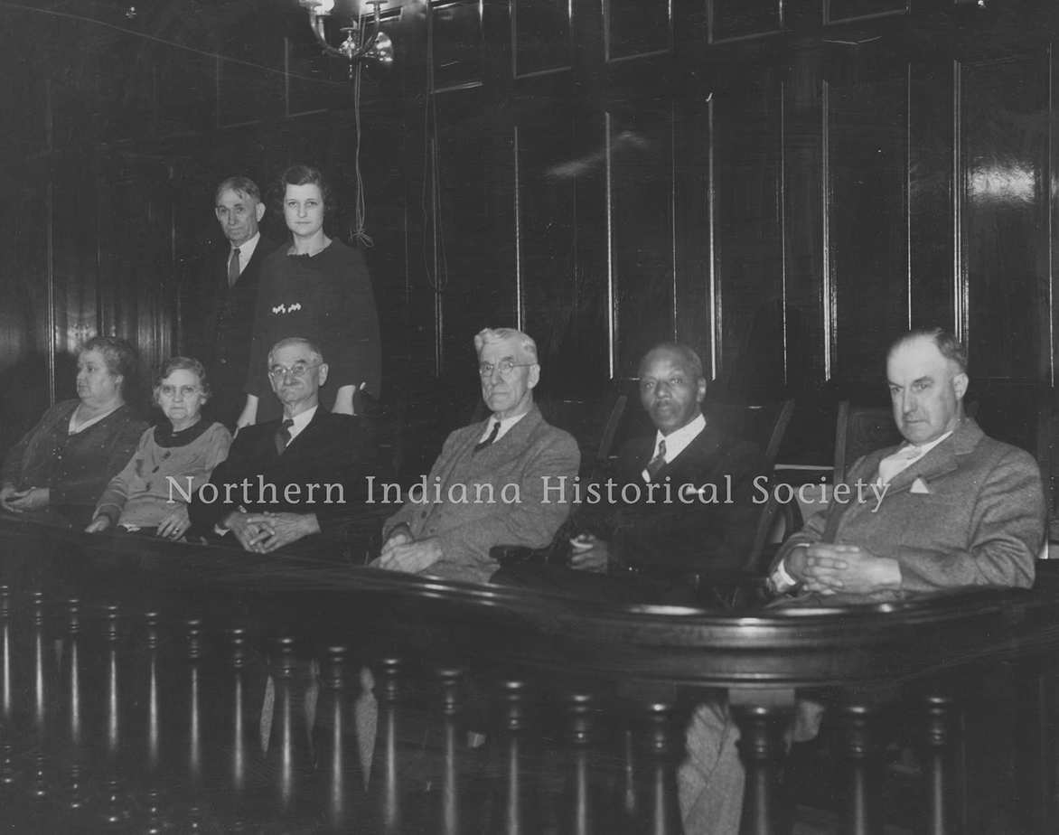 Black and white historic photo of distinguished men and women at a formal event in a wood-paneled room.