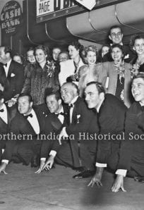 People celebrating at a historic event in a vintage setting, with men in tuxedos and women in elegant dresses.