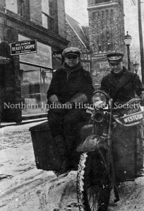 Vintage black and white photo of two men on a motorcycle outside The History Museum, snowy city streets, early 20th century urban scene, historical transportation.