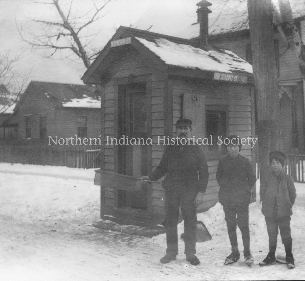 Old black and white photo of a man and two children standing outside a small historical wooden building, snow on the ground and roof.