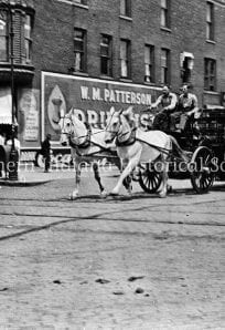 Horse-drawn carriage on a historic city street with storefronts and pedestrians, circa early 1900s.