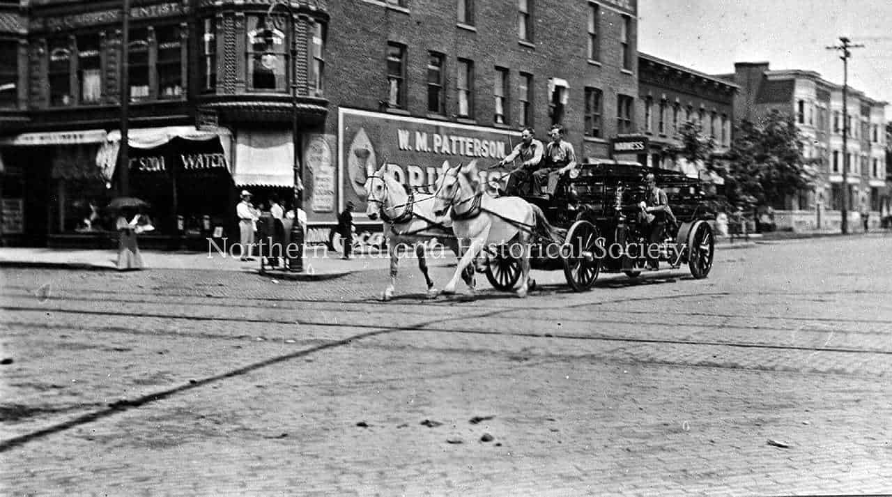 Horse-drawn carriage on a historic city street with storefronts and pedestrians, circa early 1900s.