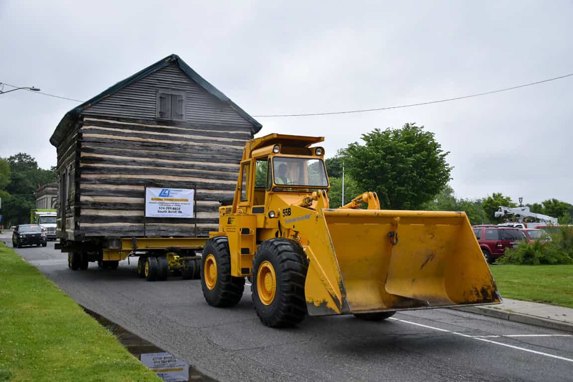Old wooden house being transported on a flatbed trailer, with a yellow front loader assisting.
