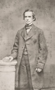 A black and white historic photo of a 19th-century man in formal attire standing beside a pedestal.
